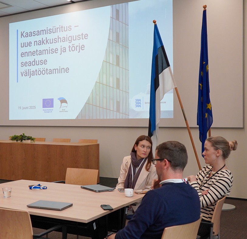 Saal, kus on presenteeritud esislaid NETS I kohtumisest, saalis istuvad lippude ees kaks naist ja mees oodates ürituse algust.